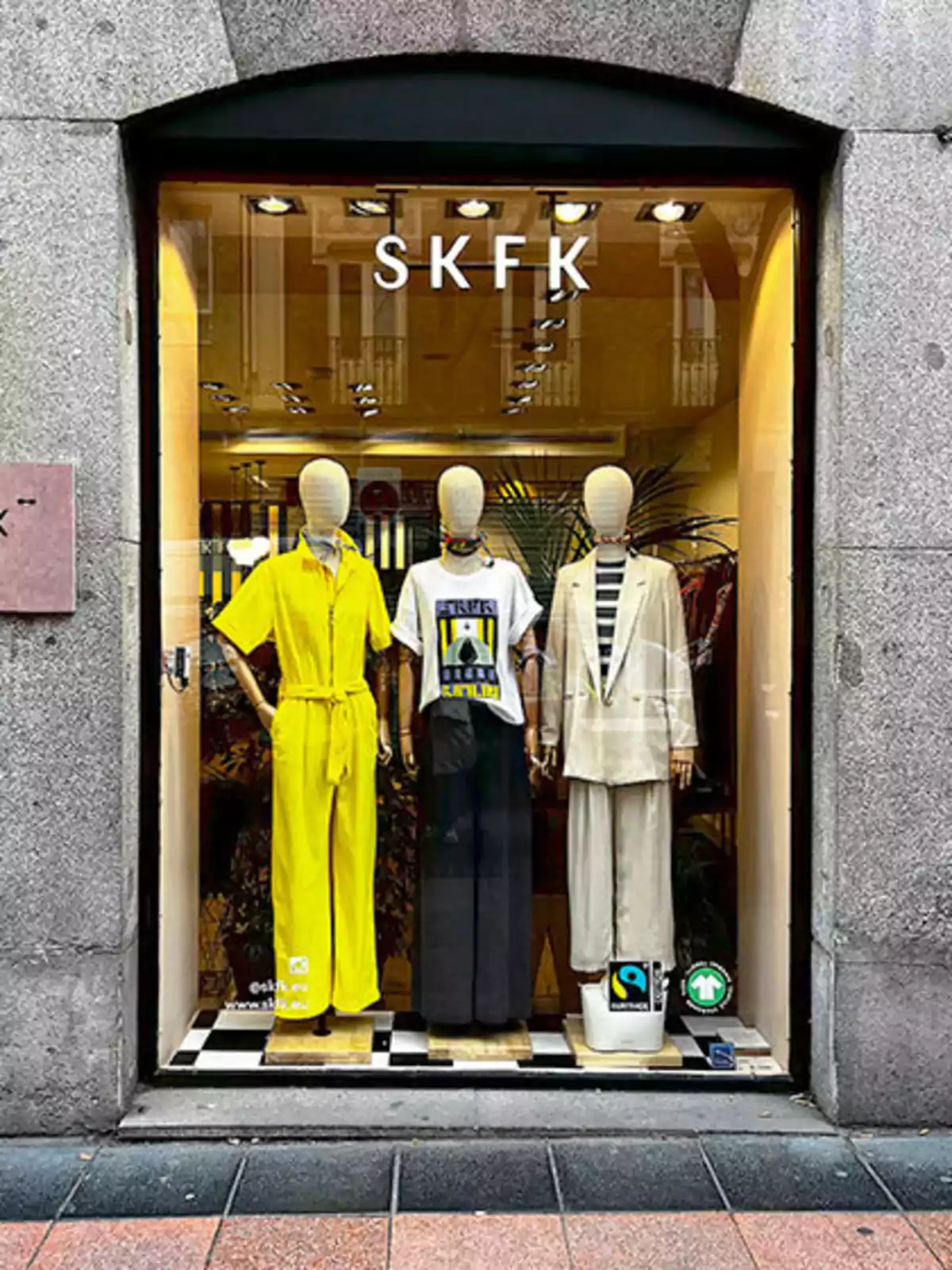 Escaparate de una tienda de ropa SKFK con tres maniquíes vistiendo diferentes conjuntos, uno amarillo, otro con camiseta blanca y pantalones oscuros, y el tercero con un traje claro, en un entorno urbano. Escaparate de una tienda de ropa SKFK con tres maniquíes vistiendo diferentes conjuntos, uno amarillo, otro con camiseta blanca y pantalones oscuros, y el tercero con un traje claro, en un entorno urbano.