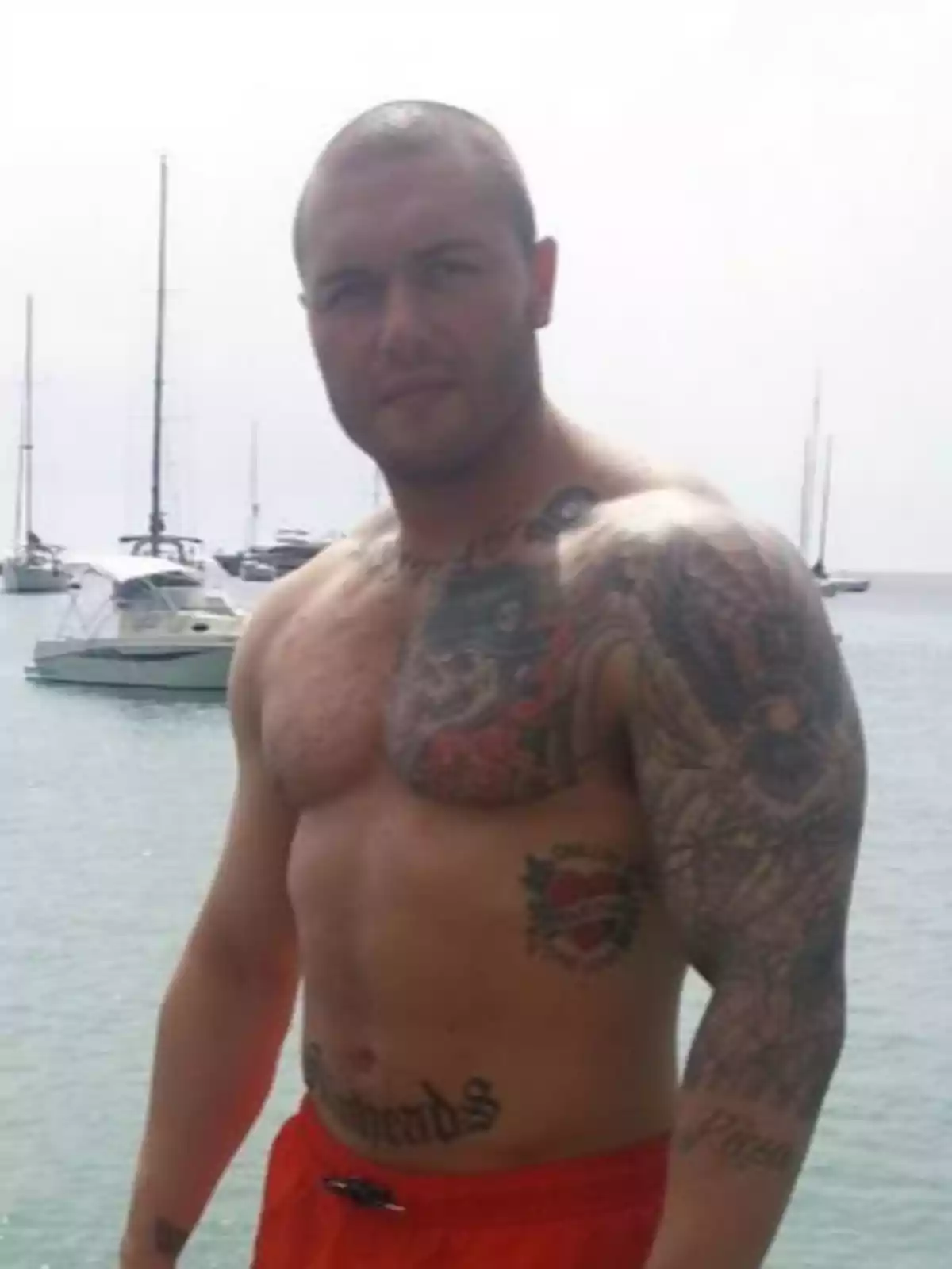 Hombre con tatuajes en el torso y brazo posando frente a un cuerpo de agua con barcos al fondo.