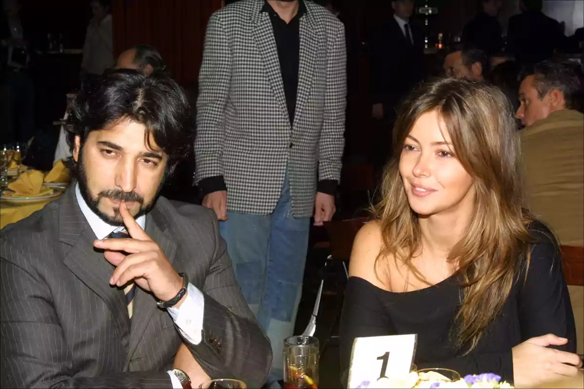 Una pareja sentada en una mesa de un restaurante, el hombre con traje y corbata y la mujer con un vestido negro, ambos parecen estar en una conversación mientras otras personas están en el fondo.