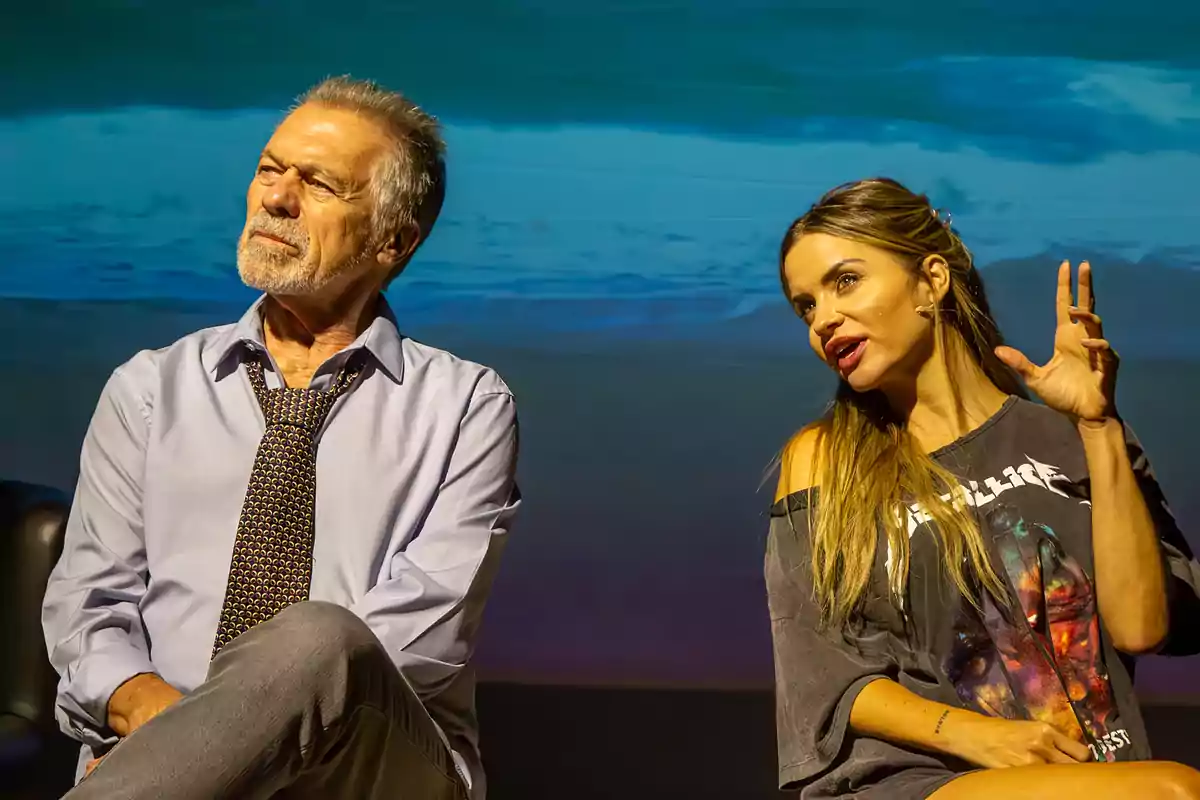 Un hombre mayor con camisa y corbata y una mujer joven con camiseta gráfica están sentados en un escenario con un fondo azul, ambos parecen estar en una conversación o presentación.