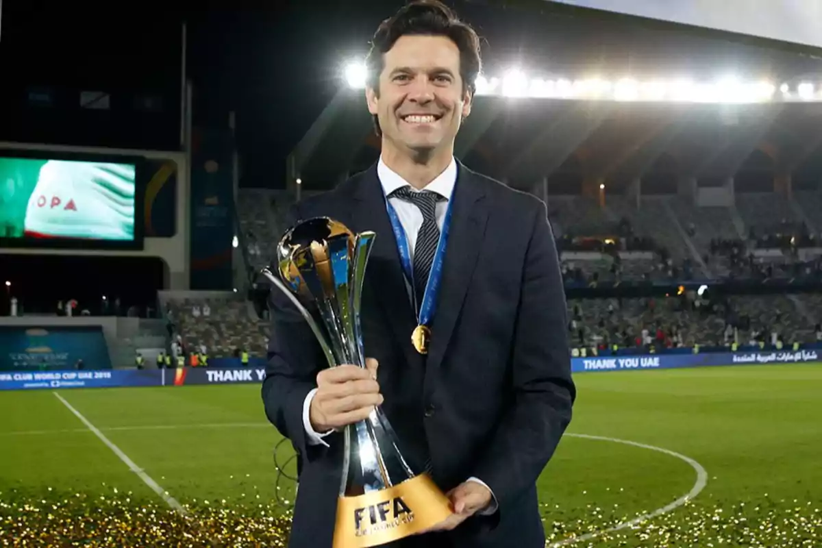 Un hombre sonriente con traje sostiene un trofeo de la FIFA en un estadio iluminado. Un hombre sonriente con traje sostiene un trofeo de la FIFA en un estadio iluminado.