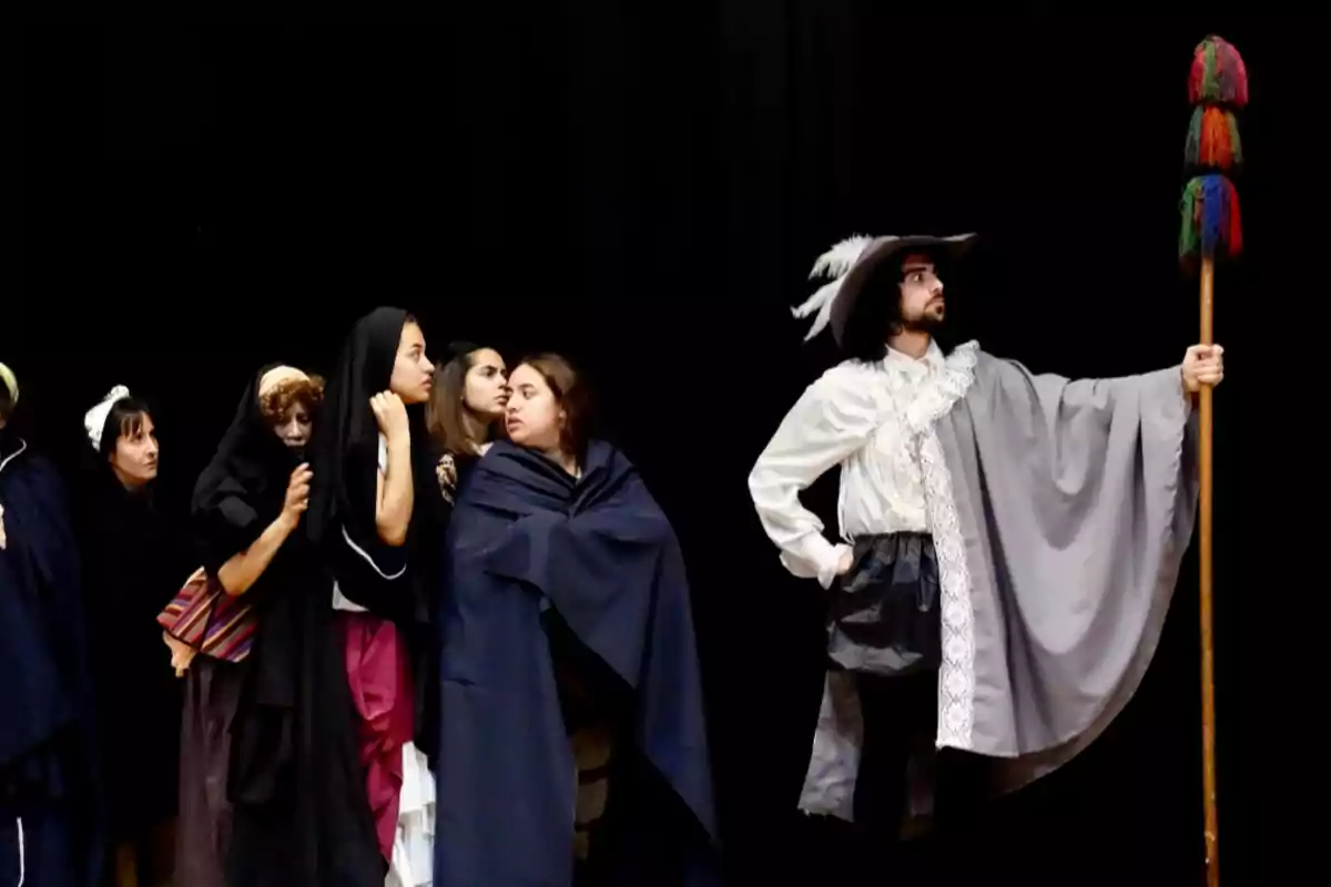 Un grupo de personas vestidas con trajes de época observa a un hombre con capa, sombrero de plumas y un bastón decorado, todos sobre un fondo negro
