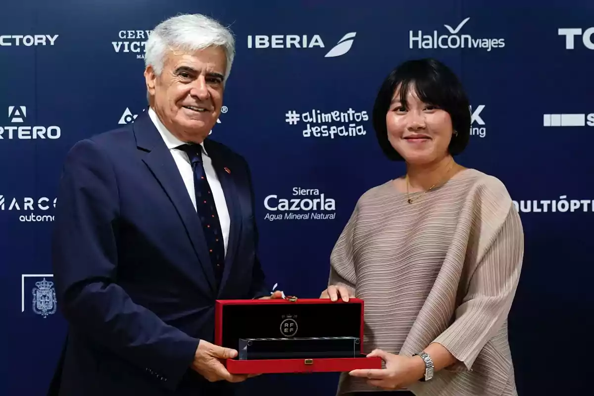 Dos personas posan para una foto mientras sostienen un estuche de presentación en un evento con un fondo lleno de logotipos de marcas. Dos personas posan para una foto mientras sostienen un estuche de presentación en un evento con un fondo lleno de logotipos de marcas.