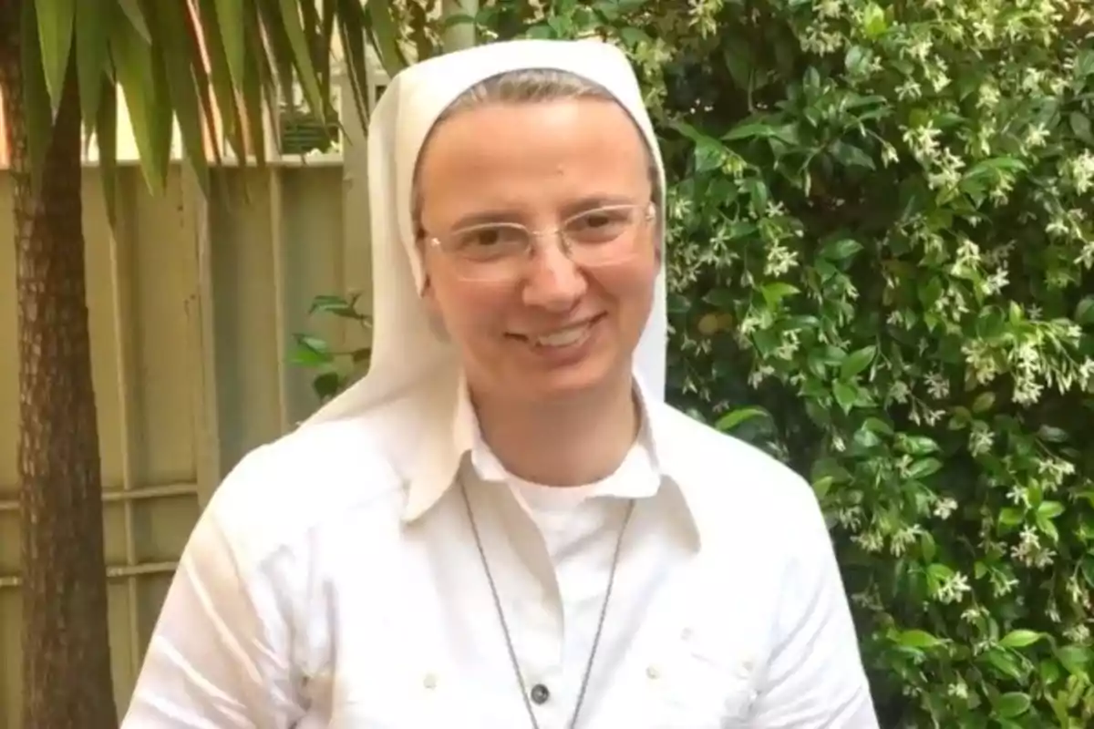 Una persona con hábito religioso y gafas sonríe frente a un fondo de plantas verdes.