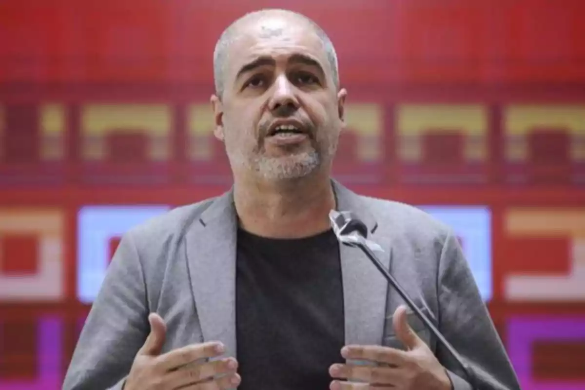Un hombre con barba y cabello corto está hablando frente a un micrófono con un fondo rojo. Un hombre con barba y cabello corto está hablando frente a un micrófono con un fondo rojo.