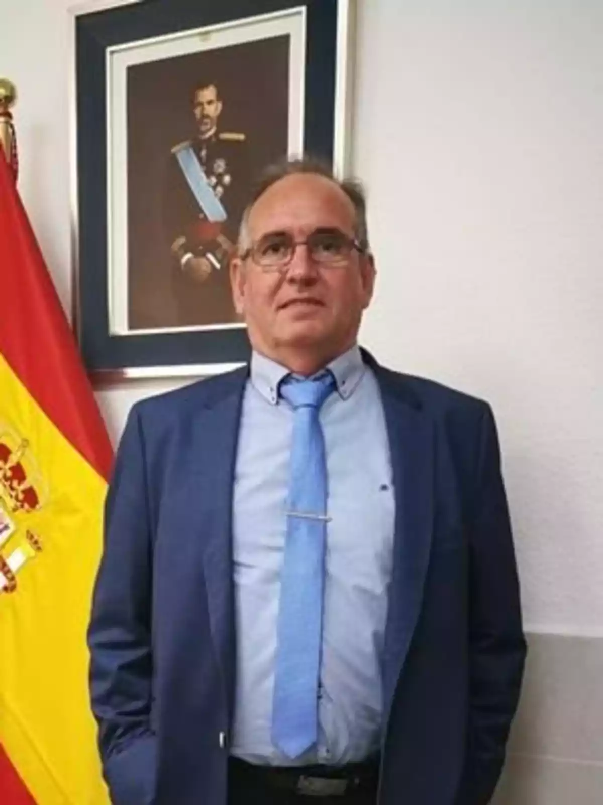 Un hombre con traje azul y corbata azul claro está de pie frente a una bandera de España y un retrato enmarcado de un hombre con uniforme militar.