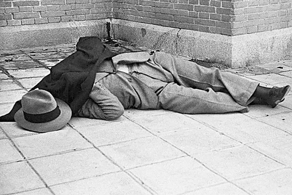 Persona acostada en el suelo con un sombrero y un abrigo en una esquina de un edificio de ladrillo.