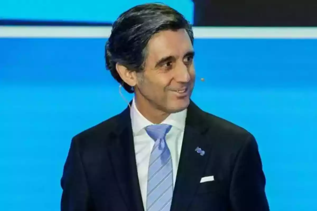 Un hombre con traje oscuro y corbata azul claro sonriendo en un escenario con fondo azul.