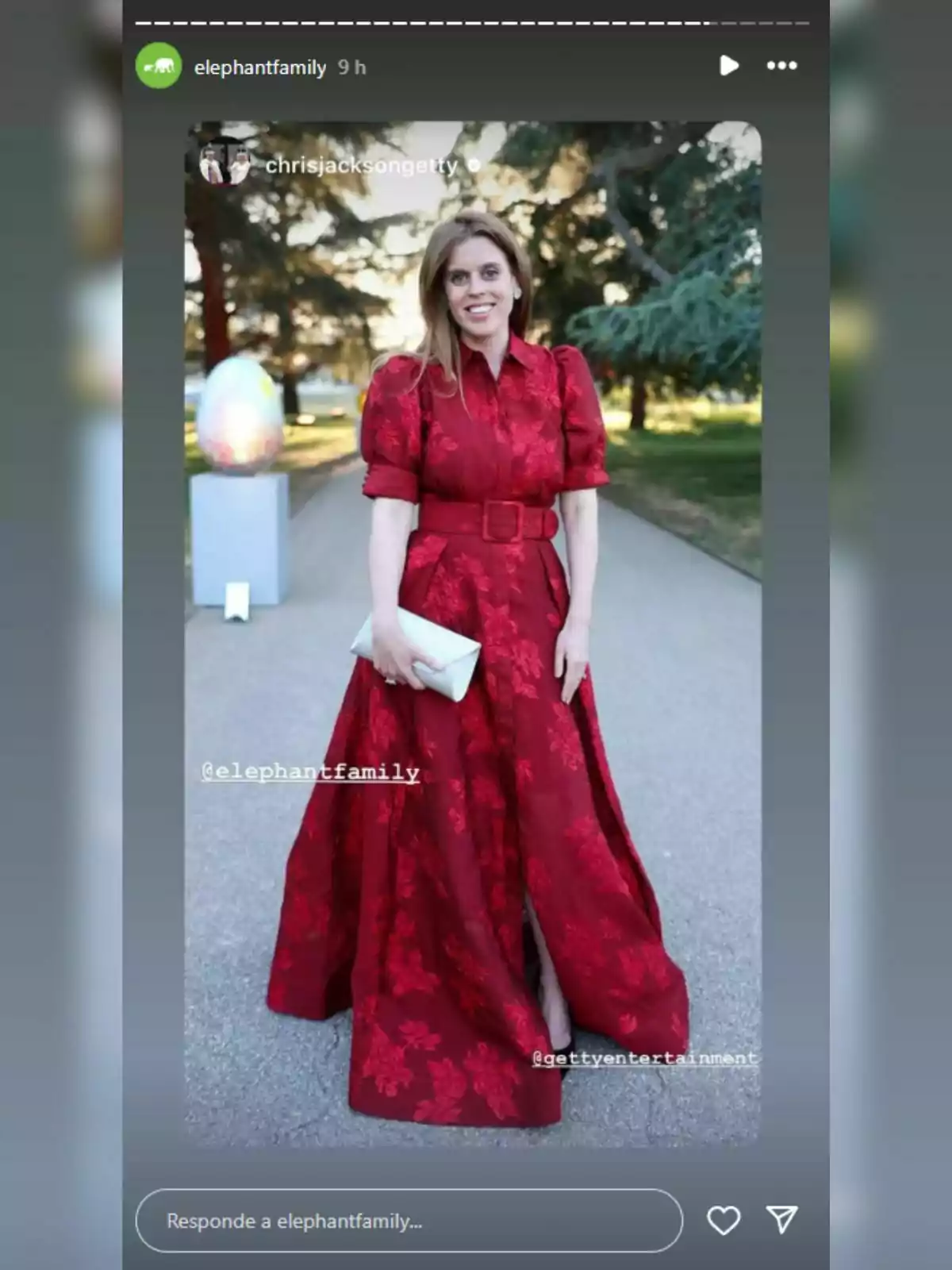 Beatriz de York con un vestido rojo elegante posa al aire libre, sosteniendo un bolso blanco, con árboles y una escultura de fondo. Beatriz de York con un vestido rojo elegante posa al aire libre, sosteniendo un bolso blanco, con árboles y una escultura de fondo.