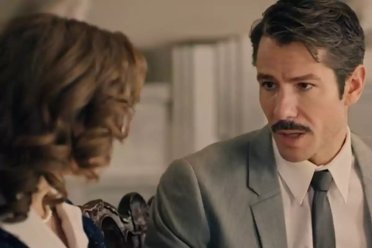 Pelayo con bigote y traje gris hablando con una mujer de cabello rizado en un ambiente interior elegante. Captura de 'Sueños de libertad' Pelayo con bigote y traje gris hablando con una mujer de cabello rizado en un ambiente interior elegante. Captura de 'Sueños de libertad'