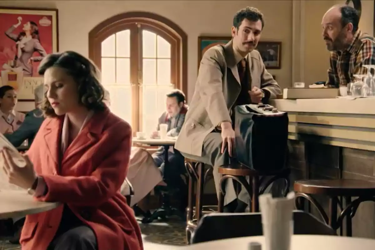 Varias personas están en una cafetería de estilo antiguo, una mujer con abrigo rojo lee un periódico en primer plano mientras un hombre con bigote y traje claro se sienta en la barra conversando con el camarero. Captura de 'sueños de libertad' Varias personas están en una cafetería de estilo antiguo, una mujer con abrigo rojo lee un periódico en primer plano mientras un hombre con bigote y traje claro se sienta en la barra conversando con el camarero. Captura de 'sueños de libertad'