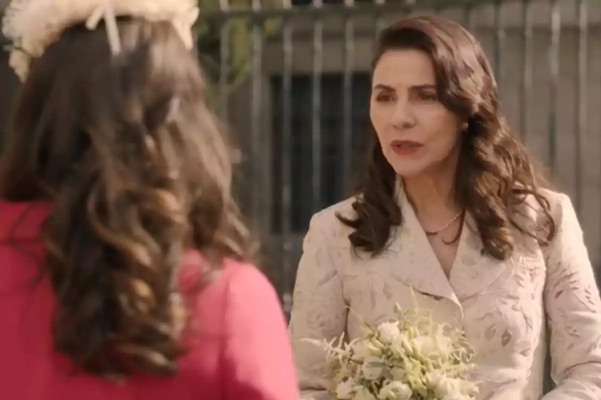 Digna y otra mujer conversan al aire libre, una de ellas sostiene un ramo de flores y viste un saco claro con bordados, mientras la otra lleva un abrigo rosa y un tocado blanco. Captura de 'Sueños de libertad' Digna y otra mujer conversan al aire libre, una de ellas sostiene un ramo de flores y viste un saco claro con bordados, mientras la otra lleva un abrigo rosa y un tocado blanco. Captura de 'Sueños de libertad'