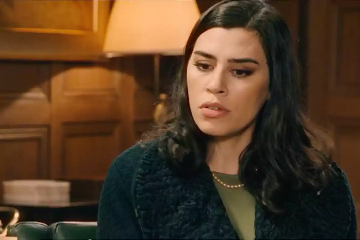 María con cabello oscuro y expresión pensativa en un ambiente interior con paredes de madera y una lámpara de fondo. Captura de 'Sueños de libertad' María con cabello oscuro y expresión pensativa en un ambiente interior con paredes de madera y una lámpara de fondo. Captura de 'Sueños de libertad'