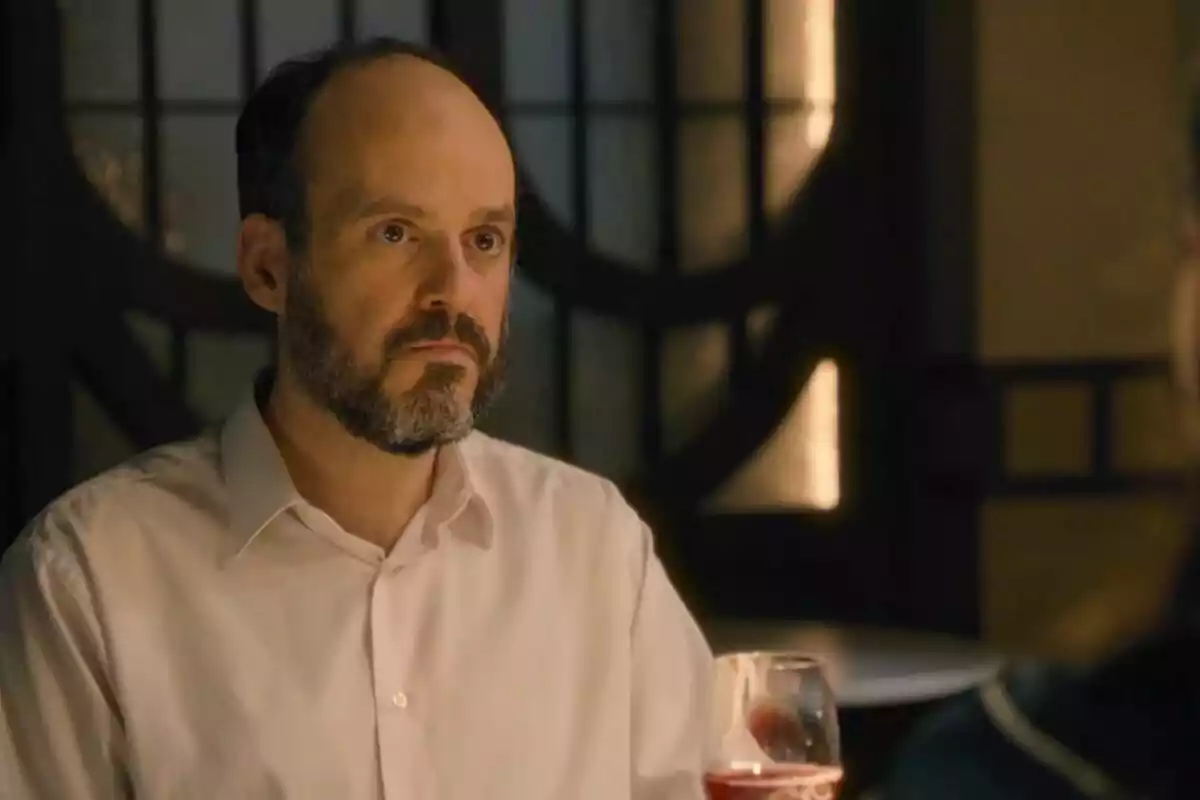 Gaspar con barba y camisa blanca está sentado en una mesa con una copa de vino frente a él, en un ambiente con iluminación cálida. Captura de 'Sueños de Libertad'