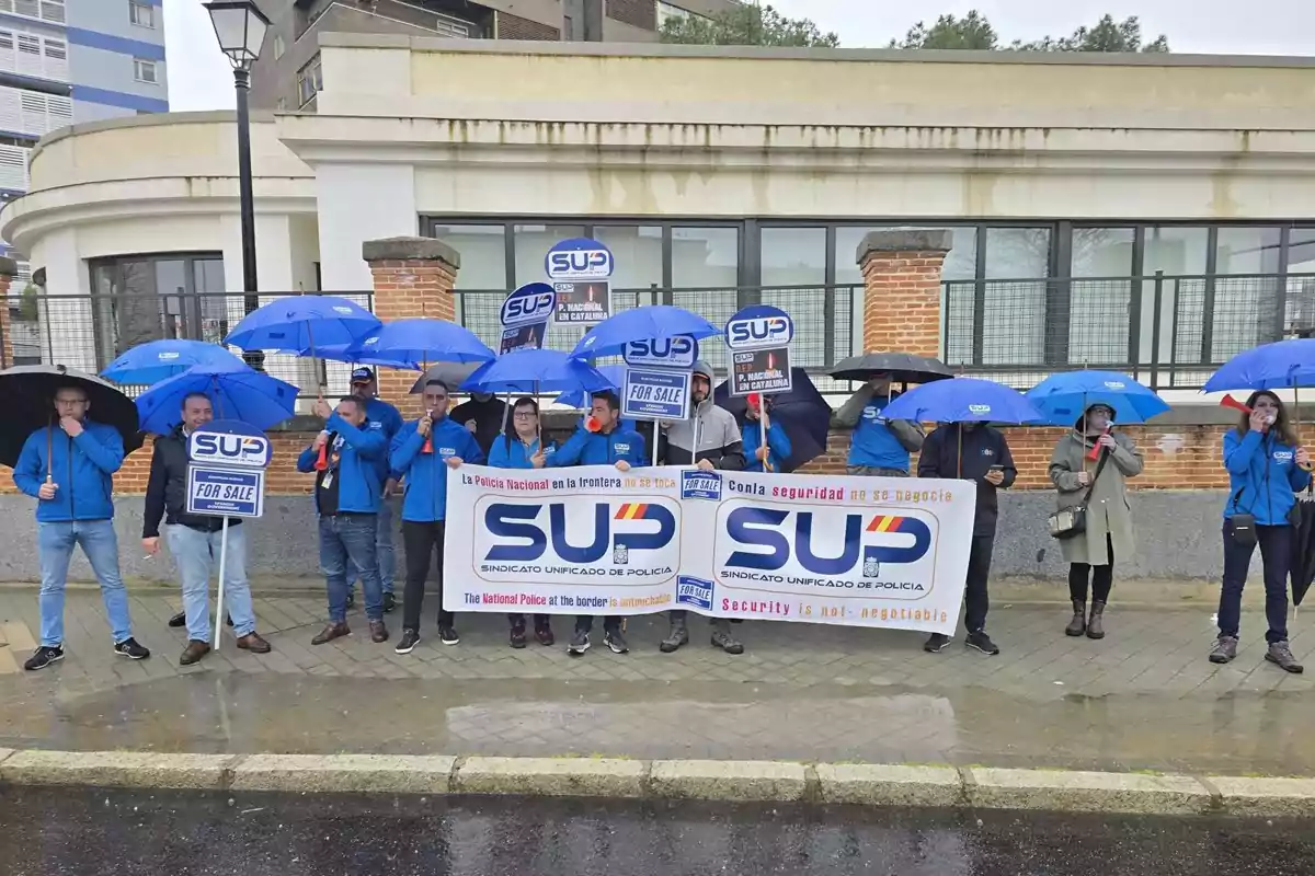 Un grupo de personas con paraguas azules sostiene pancartas y carteles del Sindicato Unificado de Policía en una manifestación bajo la lluvia frente a un edificio. Un grupo de personas con paraguas azules sostiene pancartas y carteles del Sindicato Unificado de Policía en una manifestación bajo la lluvia frente a un edificio.