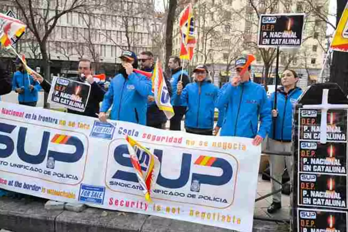 Un grupo de personas vestidas con chaquetas azules sostiene pancartas y banderas en una manifestación organizada por el Sindicato Unificado de Policía, con mensajes relacionados con la seguridad y la presencia policial en Cataluña.