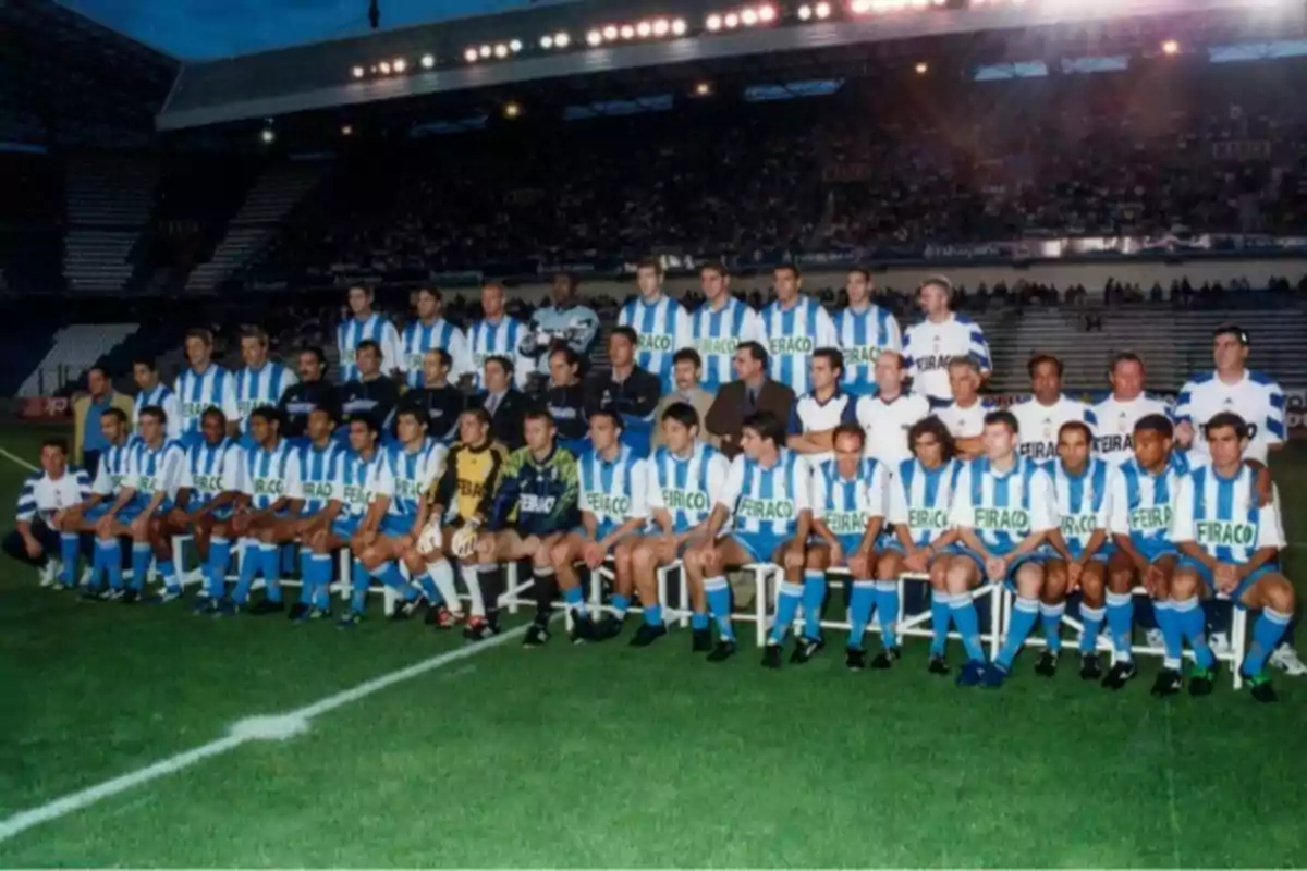 Un equipo de fútbol posando en el campo con uniformes a rayas azules y blancas, rodeados de entrenadores y personal técnico, en un estadio lleno de espectadores.