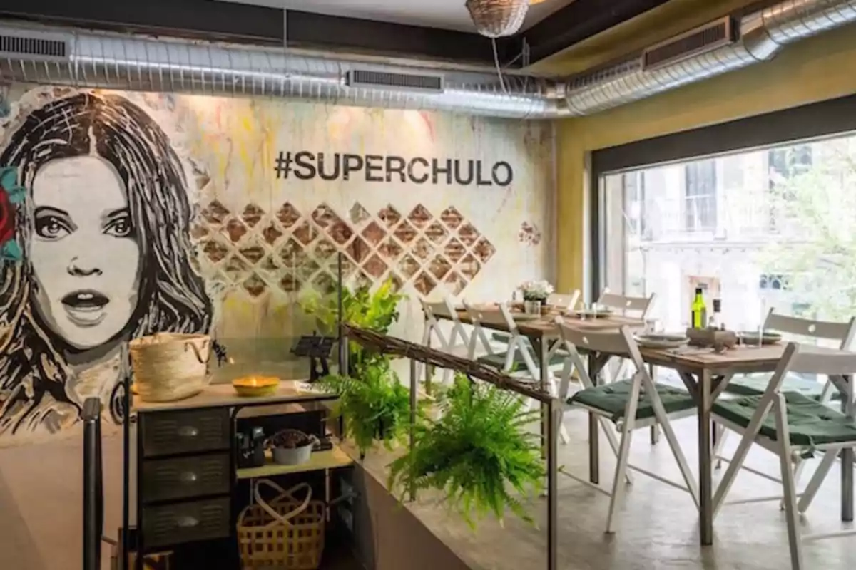 Restaurante con decoración moderna, mural de una mujer y la palabra #SUPERCHULO en la pared, mesas y sillas blancas, plantas verdes y luz natural.