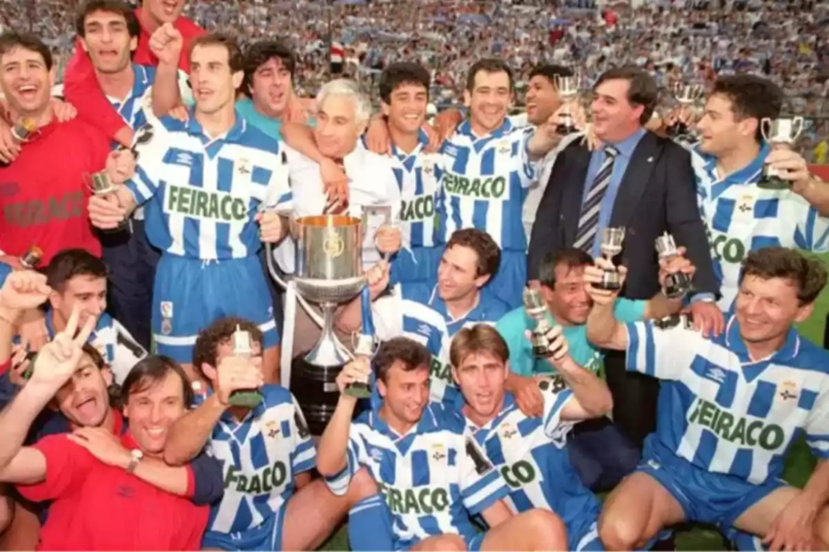 Un equipo de fútbol celebra con trofeos y una gran copa en el campo, todos visten uniformes a rayas azules y blancas y están rodeados de una multitud.