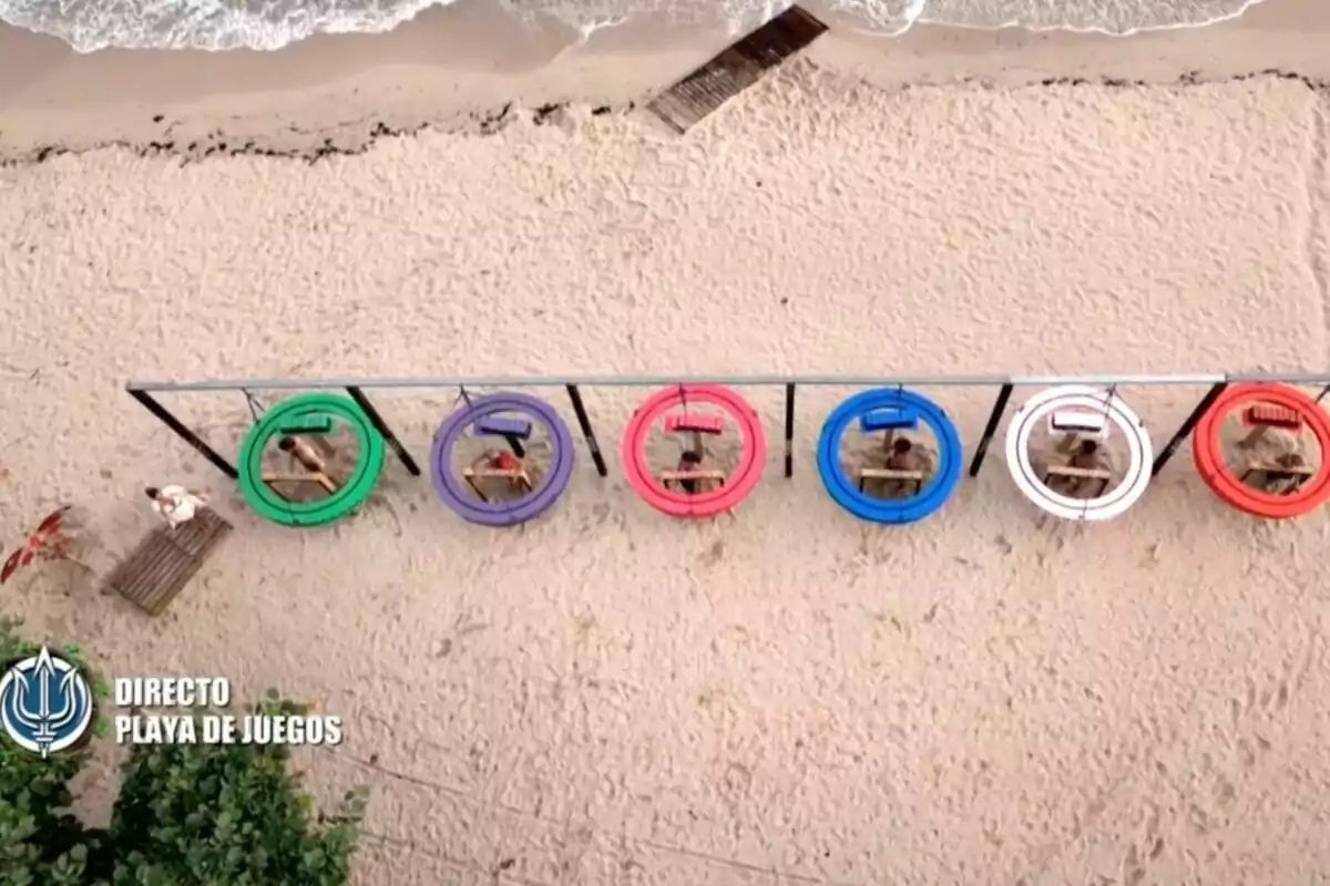 Vista aérea de una playa con seis personas sentadas dentro de aros de colores suspendidos sobre la arena frente al mar. Captura de 'Supervivientes' Vista aérea de una playa con seis personas sentadas dentro de aros de colores suspendidos sobre la arena frente al mar. Captura de 'Supervivientes'