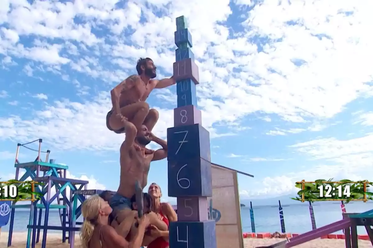 Un grupo de personas en la playa participa en un desafío de apilar bloques numerados mientras una persona se sube sobre los hombros de otra para alcanzar la cima. Captura de 'Supervivientes' Un grupo de personas en la playa participa en un desafío de apilar bloques numerados mientras una persona se sube sobre los hombros de otra para alcanzar la cima. Captura de 'Supervivientes'