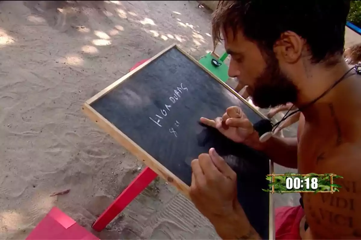 Montoya escribe en una pizarra pequeña mientras está sentado en la arena con un cronómetro en la esquina inferior derecha mostrando 00:18. Captura de 'Supervivientes' Montoya escribe en una pizarra pequeña mientras está sentado en la arena con un cronómetro en la esquina inferior derecha mostrando 00:18. Captura de 'Supervivientes'