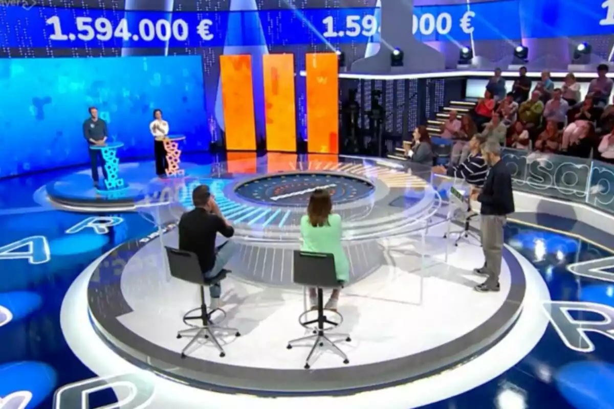 Un programa de concurso en un estudio de televisión con participantes de pie detrás de podios iluminados y un público observando desde las gradas. Captura de 'Pasapalabra' Un programa de concurso en un estudio de televisión con participantes de pie detrás de podios iluminados y un público observando desde las gradas. Captura de 'Pasapalabra'