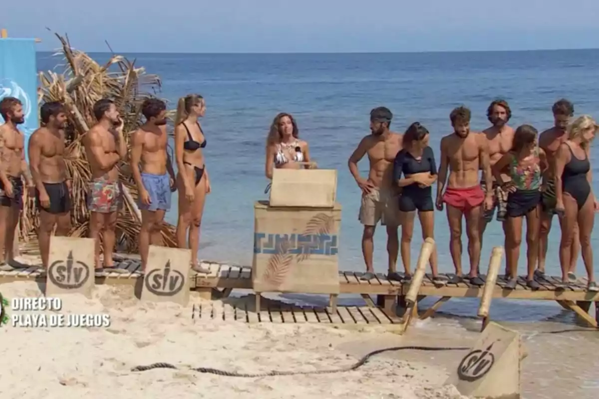 Un grupo de personas en traje de baño está reunido en una playa junto a un podio de madera, con el mar de fondo y un cartel azul a la izquierda. Captura de 'Supervivientes' Un grupo de personas en traje de baño está reunido en una playa junto a un podio de madera, con el mar de fondo y un cartel azul a la izquierda. Captura de 'Supervivientes'