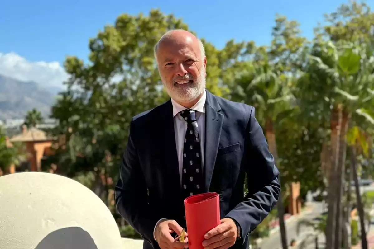 Un hombre con traje oscuro y corbata sostiene un objeto rojo en un entorno exterior con árboles y cielo azul.