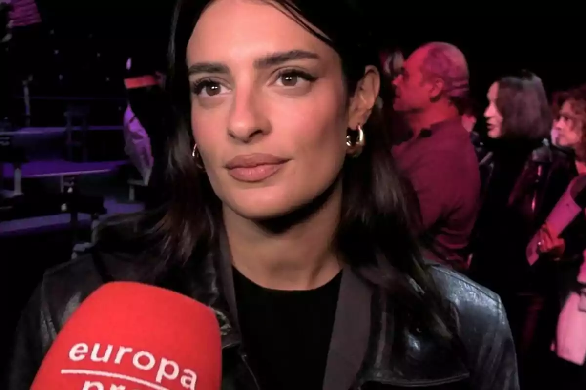 Plano corto de Susana Bicho con chaqueta de cuero es entrevistada por un micrófono rojo en un evento con varias personas al fondo. Plano corto de Susana Bicho con chaqueta de cuero es entrevistada por un micrófono rojo en un evento con varias personas al fondo.