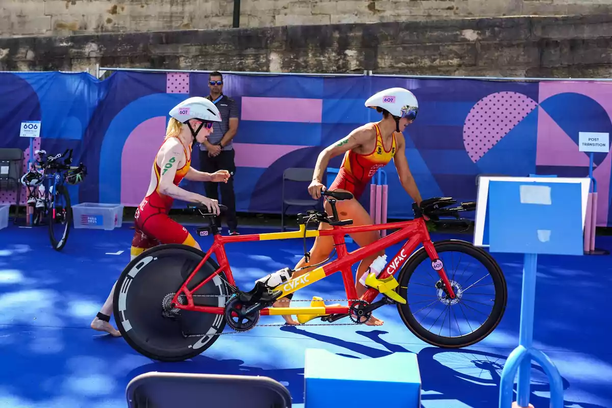 Dos atletas con uniformes rojos y amarillos se preparan para montar una bicicleta tándem en una zona de transición durante una competencia deportiva.