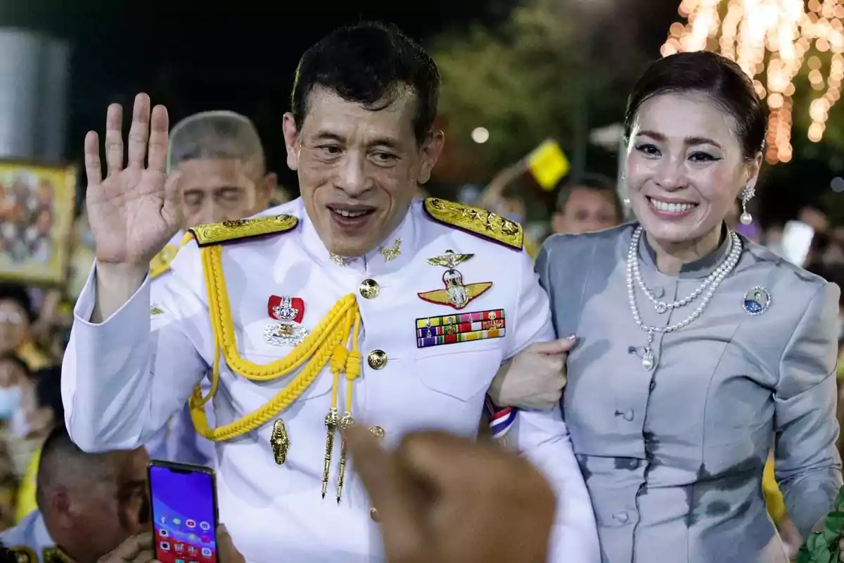 Un hombre y una mujer sonrientes, ambos vestidos con trajes formales, rodeados de personas y luces de fondo.