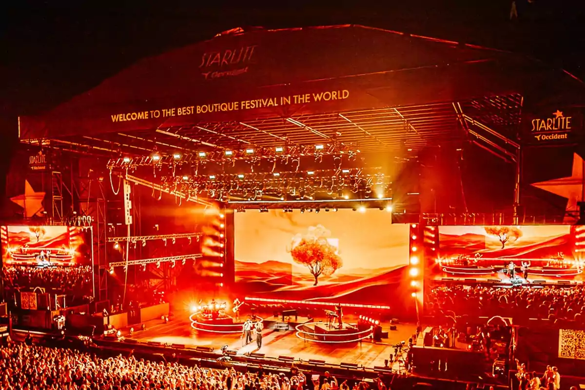 Escenario iluminado con luces naranjas y rojas durante un festival de música con una gran multitud de personas.