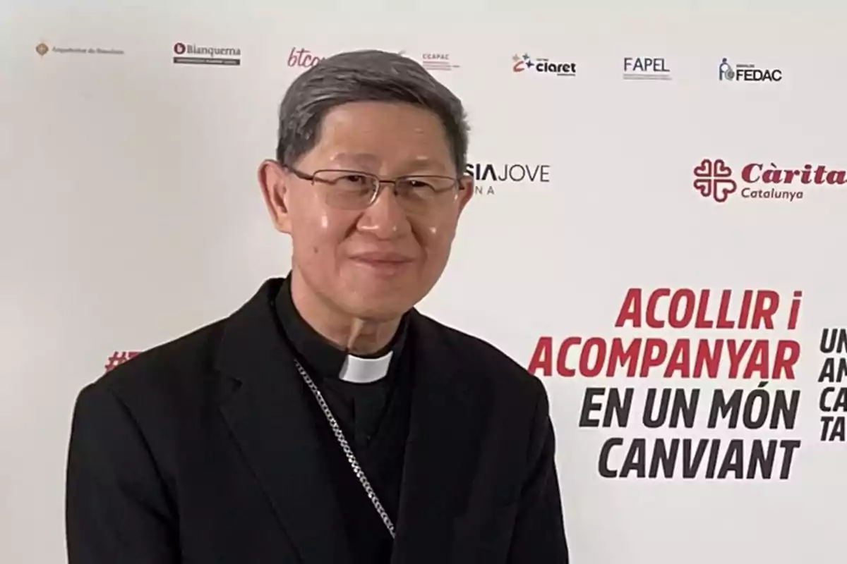 Un hombre con gafas y vestimenta clerical frente a un fondo con logotipos y texto en catalán.