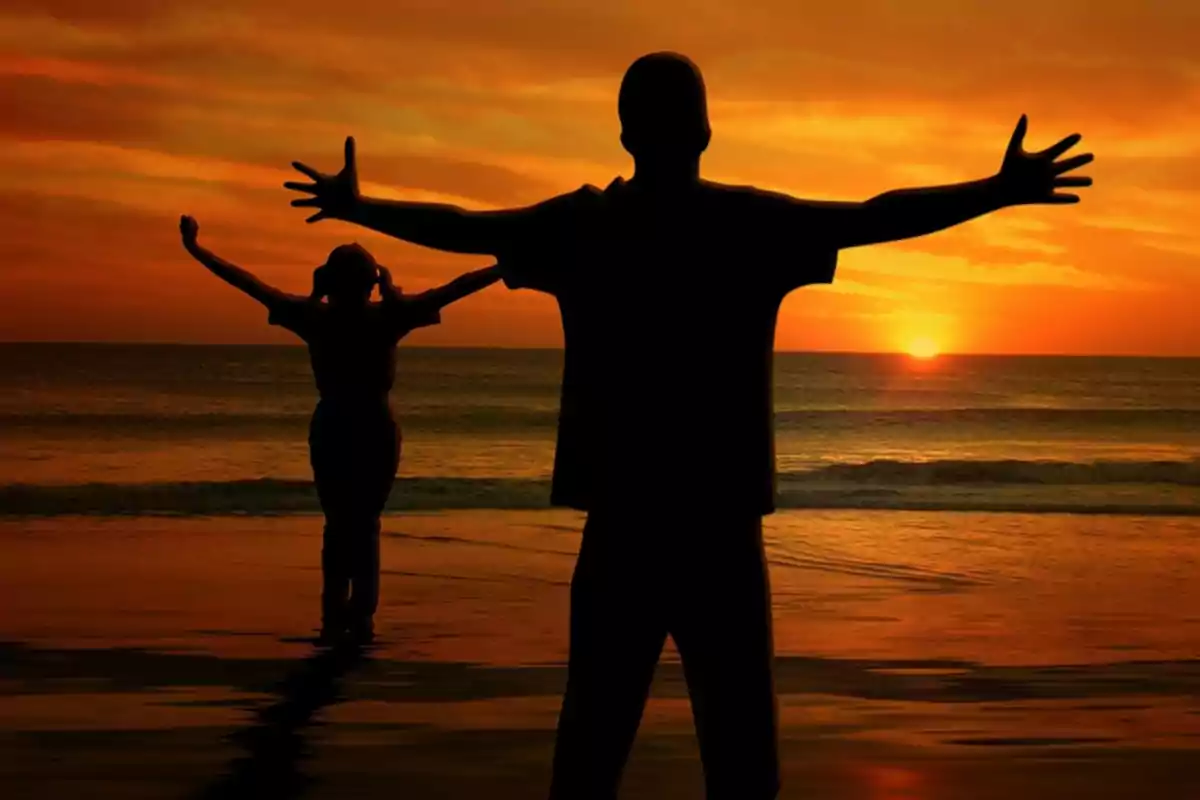 Siluetas de dos personas con los brazos extendidos frente a un atardecer en la playa.