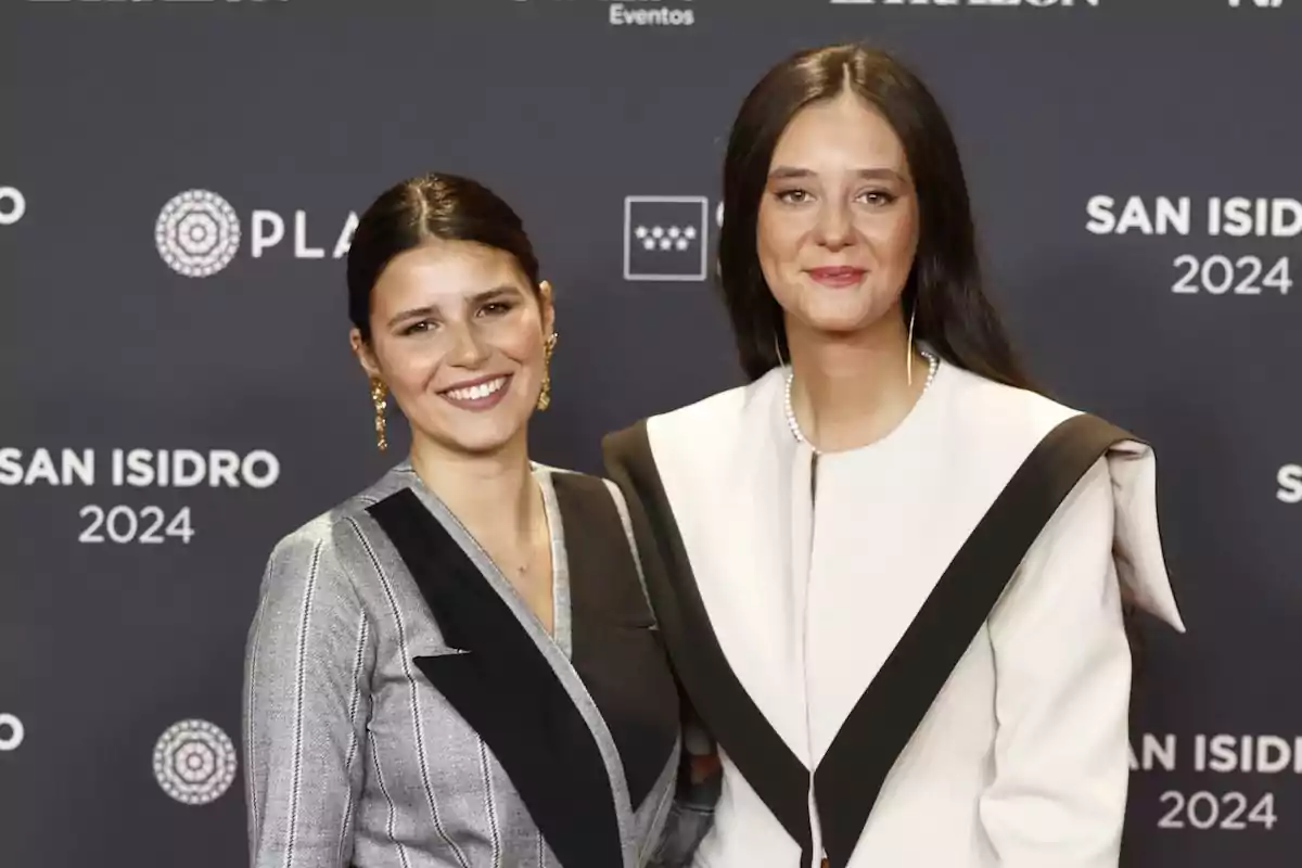 Dos mujeres posan sonrientes en un evento de San Isidro 2024, vestidas con elegantes trajes en tonos blanco y negro.