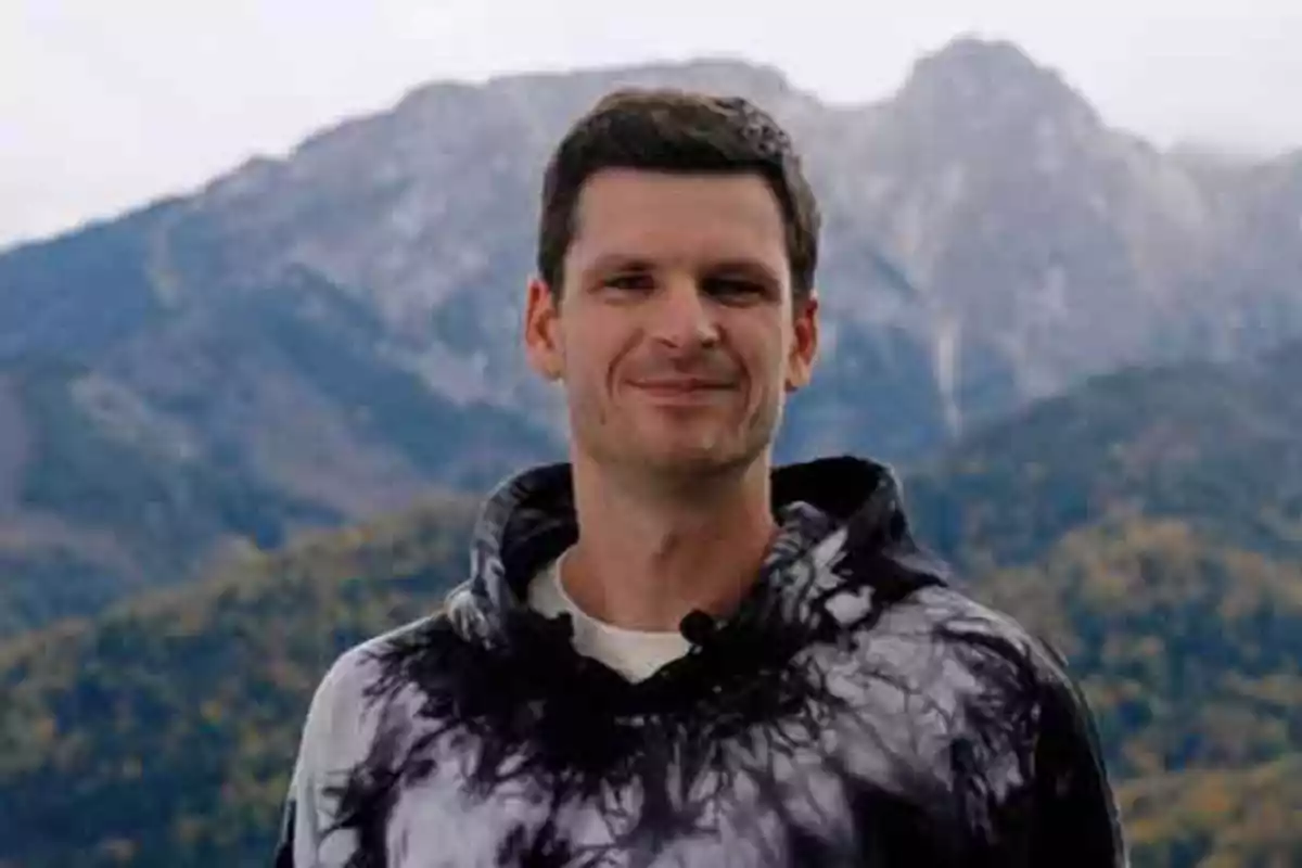 Un hombre con sudadera de diseño abstracto sonríe frente a un paisaje montañoso.