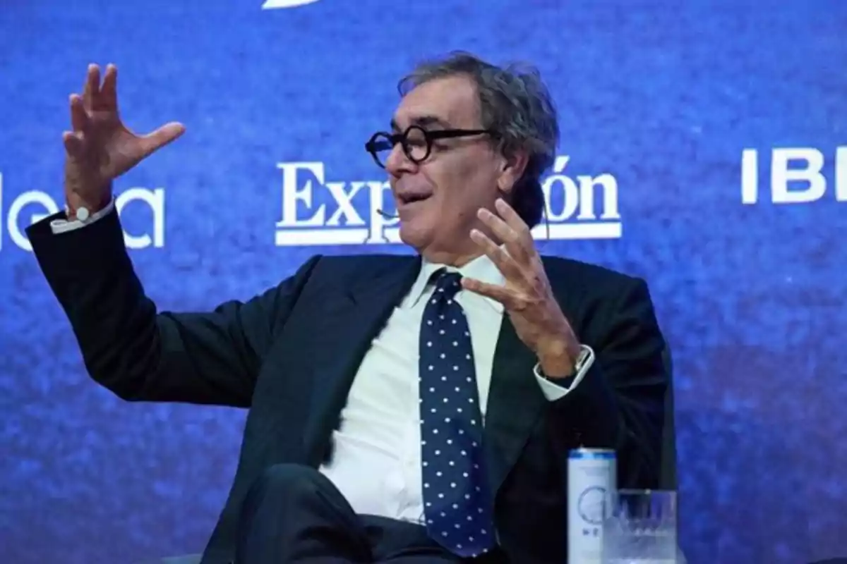 Hombre de traje y corbata con gafas hablando y gesticulando con las manos frente a un fondo azul con logotipos de empresas Hombre de traje y corbata con gafas hablando y gesticulando con las manos frente a un fondo azul con logotipos de empresas