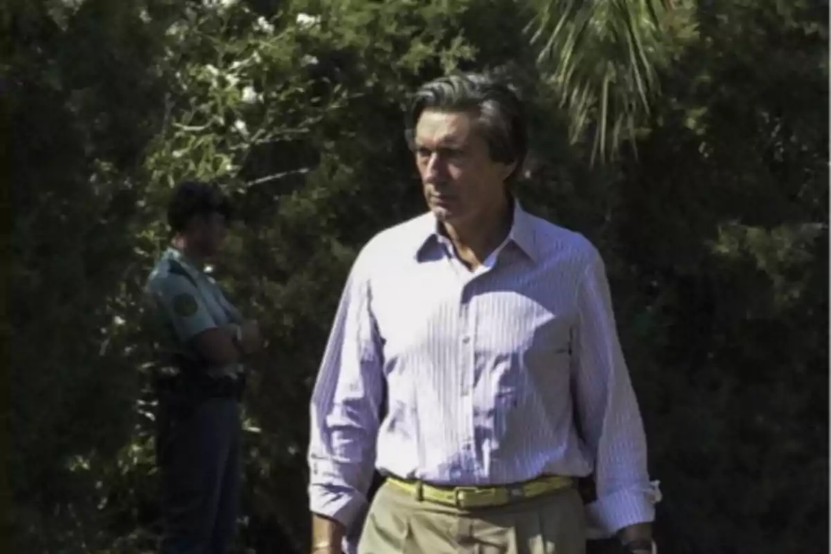 Un hombre de cabello oscuro y camisa a rayas camina al aire libre con un guardia de seguridad al fondo entre árboles.