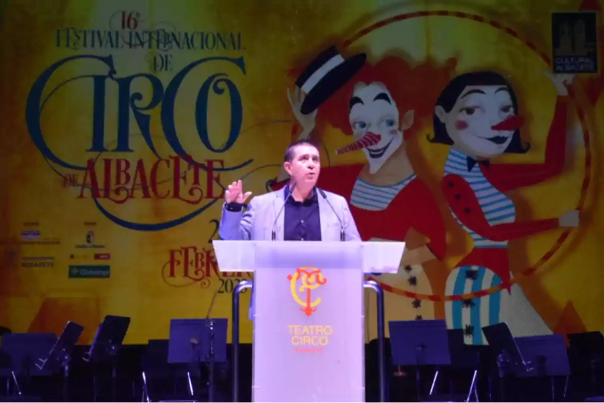 Un hombre hablando en un podio frente a un colorido cartel del Festival Internacional de Circo de Albacete.