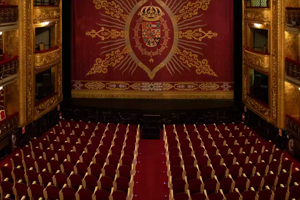 Un teatro elegante con asientos rojos y un telón decorado con un escudo y detalles dorados. Un teatro elegante con asientos rojos y un telón decorado con un escudo y detalles dorados.