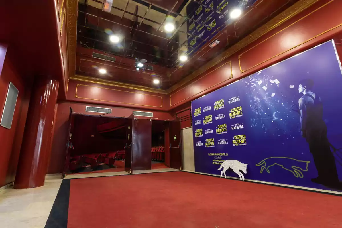 Vestíbulo de un teatro con paredes y techo rojos, una alfombra roja y un cartel promocional de una obra llamada 