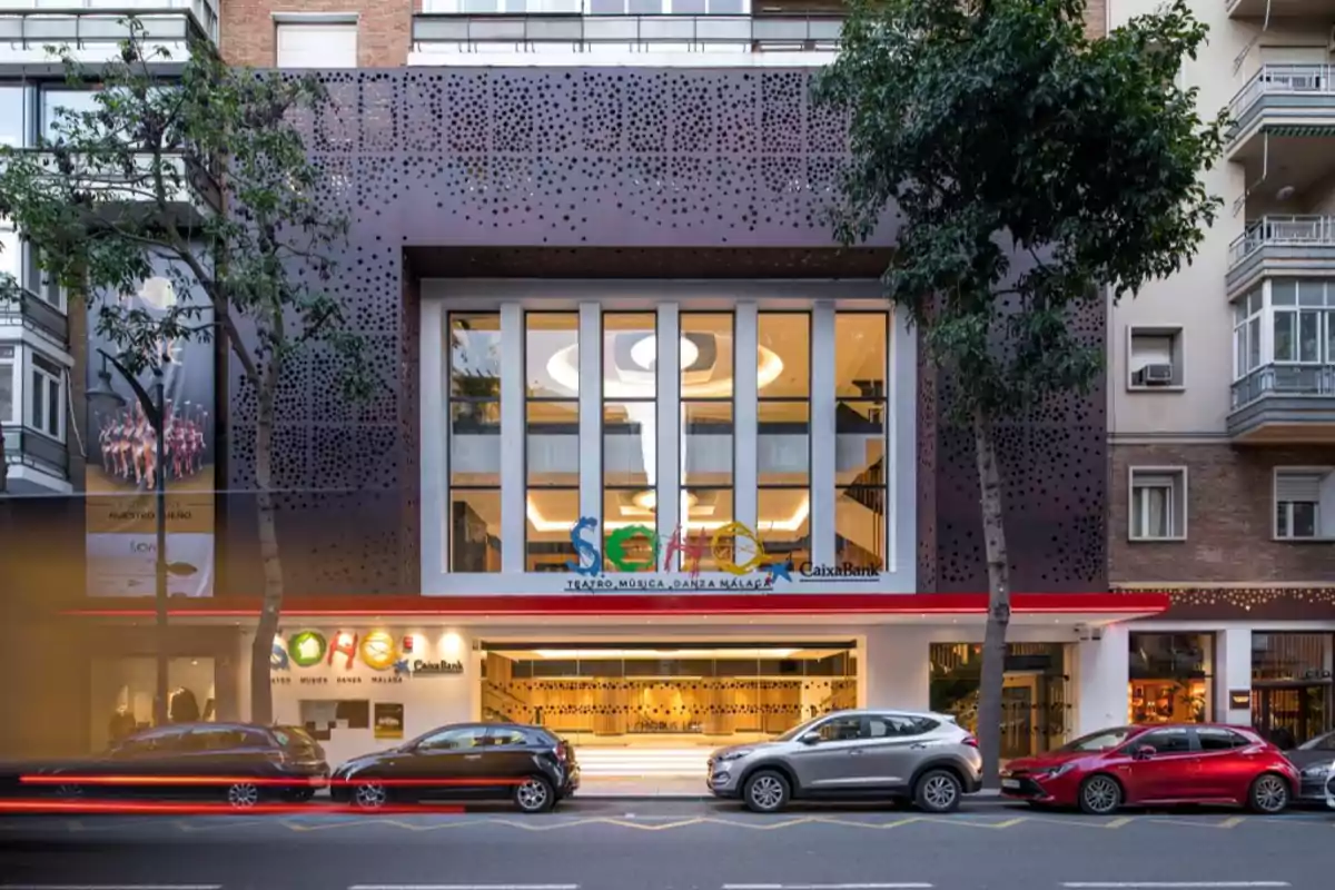 Fachada de un teatro moderno con coches estacionados en la calle frente a él.