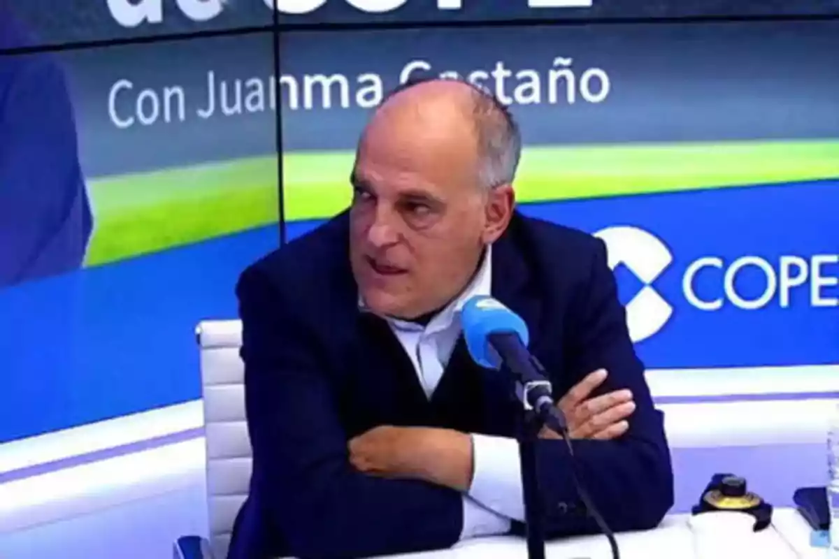 Hombre calvo con chaqueta oscura hablando en un estudio de radio con un micrófono azul.