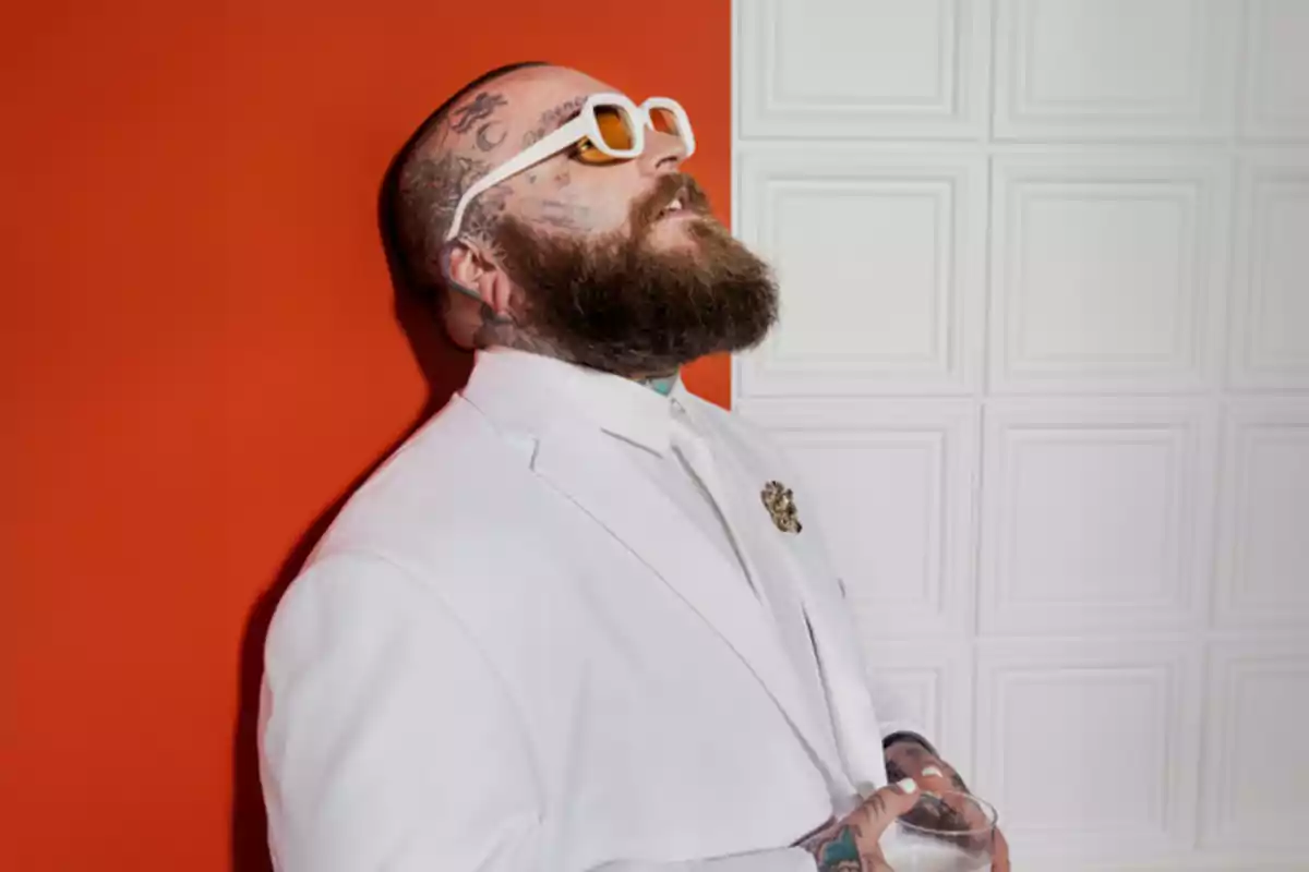 Hombre con barba y tatuajes usando gafas de sol y traje blanco, apoyado en una pared naranja.