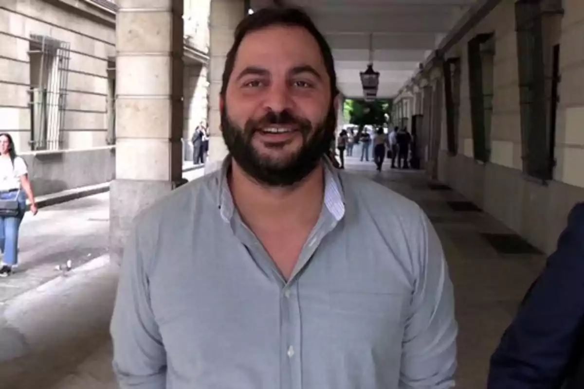 Un hombre con barba y camisa clara sonríe mientras camina por un pasillo exterior con columnas y personas al fondo.