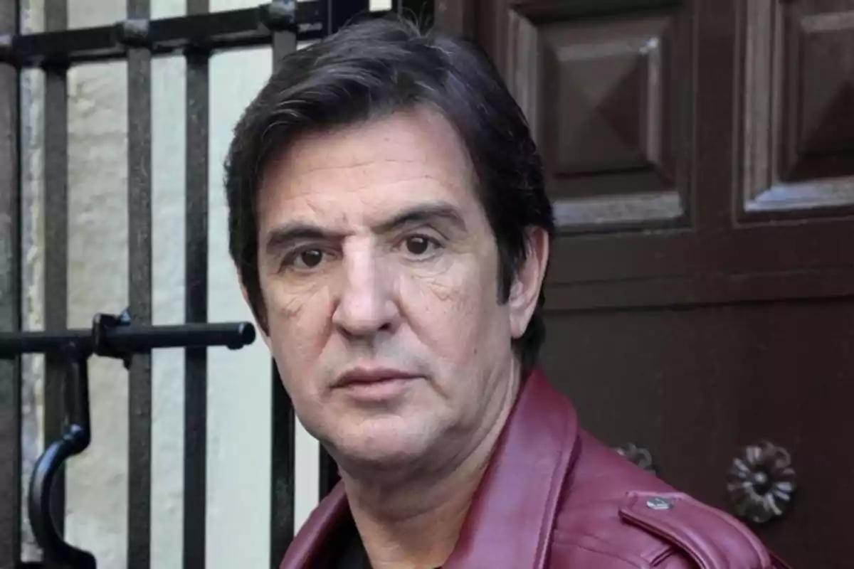 Un hombre con chaqueta de cuero roja frente a una puerta de madera y una reja de metal. Un hombre con chaqueta de cuero roja frente a una puerta de madera y una reja de metal.