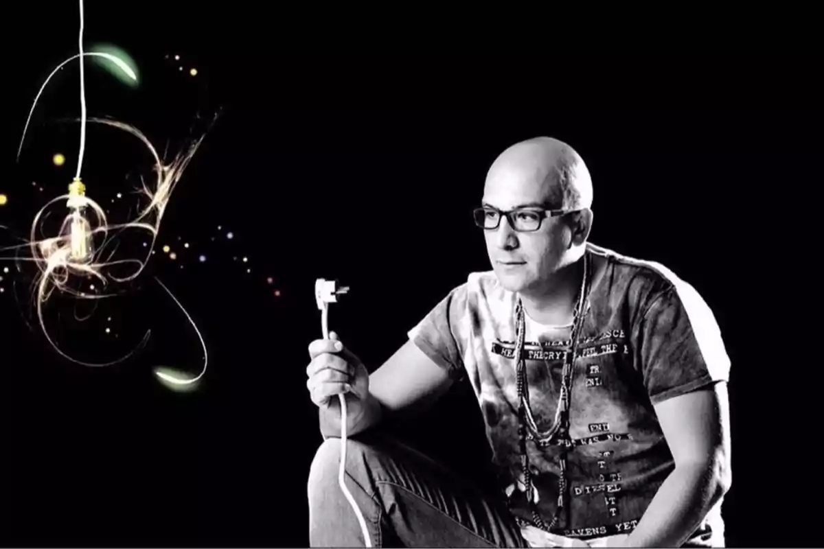 Un hombre con gafas sostiene un cable mientras observa una bombilla colgante rodeada de efectos luminosos en un fondo negro. Un hombre con gafas sostiene un cable mientras observa una bombilla colgante rodeada de efectos luminosos en un fondo negro.