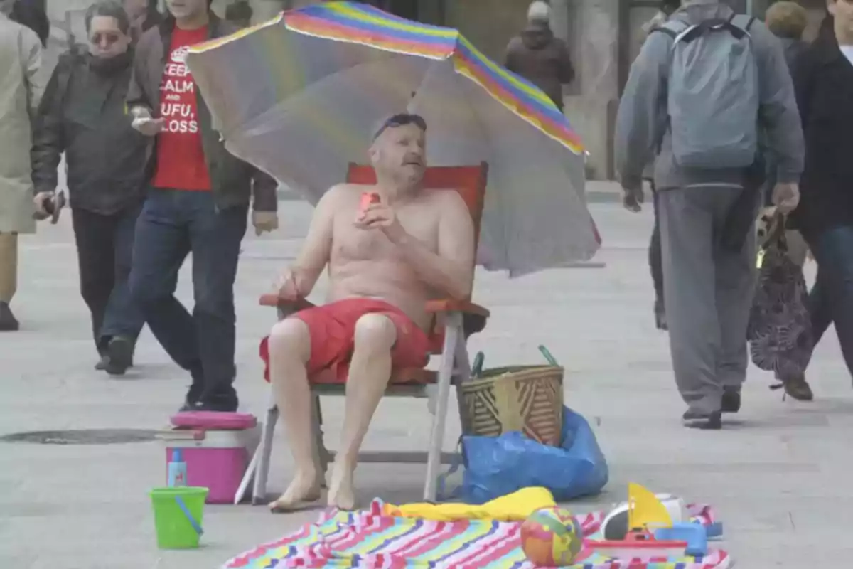 Un hombre en traje de baño rojo está sentado en una silla de playa bajo una sombrilla multicolor en medio de una calle concurrida, rodeado de objetos de playa como una toalla, un balde y una pelota.