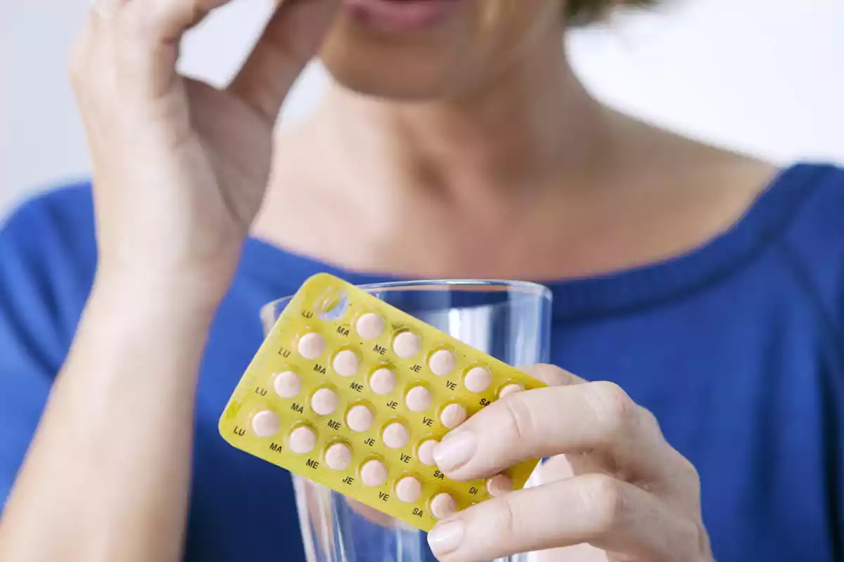 Persona tomando una pastilla de un blíster amarillo mientras sostiene un vaso de agua.